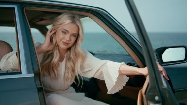 Güzel okyanus arkaplanındaki otomobil ön koltuğunda poz veren sevimli sarışın. Retro araba görünümlü kamerada poz veren kaygısız kadın. Çekici genç bayan gezgin inanılmaz deniz kıyısına araba yolculuğunun tadını çıkarıyor.