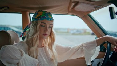 Araba kuaföründe oturan kız direksiyona el koyup bandana takıyor. Güzel kadın hipster rahat bir retro otomobilde rahatlamayı sever. Otomatik olarak poz veren çekici tasasız model.