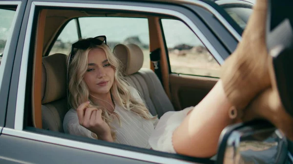 Gorgeous model posing in automobile putting feet out open window close up. Beautiful woman traveler sitting in front passenger seat retro car with sunglasses on head. Carefree blonde relaxing in auto.