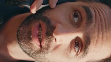 İspanyol adam güneş ışığında cep telefonunu arıyor. Doğanın dikey videosu. Yakın çekim. Rahatlamış gülümseyen turist yaz parkında cep telefonuyla konuşuyor. Şehir kafeteryasında akıllı telefonla konuşan mutlu yetişkin adam.