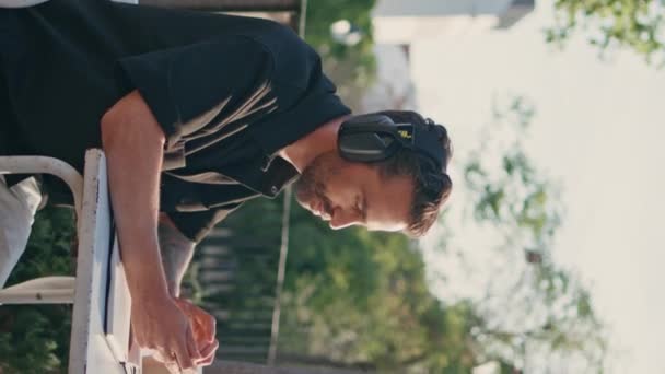受到启发的男人在耳机里欣赏着最喜爱的音乐 坐在城市街道的垂直天使的近旁 意大利自由职业者夏天拿着智能手机听着有节奏的歌曲 布鲁内特的家伙用手机 — 图库视频影像