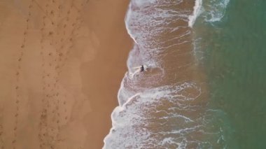 Altın kumsalda elinde sörf tahtasıyla yürüyen bir sörfçü. Tanınmayan kişi sörf yaptıktan sonra su sıçratıyor. Turkuaz okyanus suyu süper yavaş çekimde manzaralı kıyı şeridine akıyor..