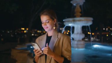 Akşam şehrinde gülümseyen kadın cep telefonunu kaydırıyor. Nezarethanedeki zarif kadın aydınlanmış su çeşmesinde arkadaşlarını bekliyor. Dışarıda akıllı telefonları dinleyen güzel bir kız. Gece hayatı konsepti
