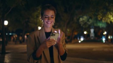 Akıllı telefondan mesaj atan şehirli bir kadın gece sokağa yalnız çıkıyor. Gülümseyen kadın sosyal medyada sokak mesajlaşması yapıyor. Çekici iş kadını görünümlü cep telefonu ekranı tarama çevrimiçi alıkoyma aygıtı.