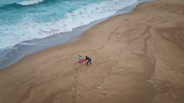 Drone manzaralı sörfçü kumlu sahilde tek başına egzersiz yapıyor. Sörfçü, ıslak kumda, teknenin yanında cesedi ısıtıyor. Sahili kaplayan köpüklü okyanus suyu. Resimli deniz doğası ağır çekim. Etkin hobi kavramı