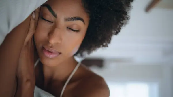Serene woman sleeping pillow at hotel closeup. Curly hair lazy girl opening eyes looking camera at bedroom portrait. African american model napping bed resting at morning alone. Calm lady awake indoor