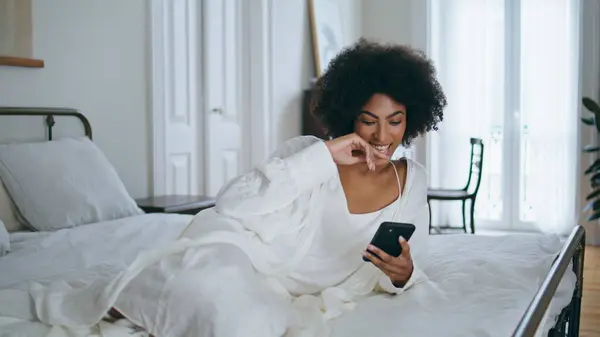 Laughing model watching phone funny video at home. Relaxed african lady laying bed holding smartphone device. Cheerful girl surfing cellphone app at white bedroom. Domestic weekend lifestyle concept