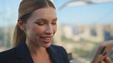 Blue Sky 'da akıllı telefonları dinleyen mutlu bir kadın. Gülümseyen menajer şehir terasında hücre izleyerek sosyal medyayı kullanıyor. Çekici iş kadını, modern cep telefonunu kaydırıyor ve tatilin tadını çıkarıyor..