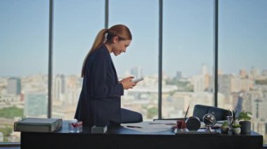 Cam ofiste akıllı telefonlara bakan kaygısız bir işçi. Gülümseyen iş kadını dinleniyor cep telefonu mesajları çevrimiçi. Rahat kadın yönetici çıplak ayakla panoramik pencerede oturuyor.