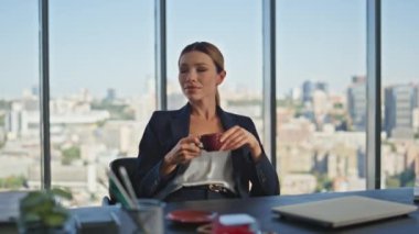 Sakin müdür modern ofiste kahve içiyor. Gülümseyen kadın panoramik şehir manzarasında içeceğini yudumlarken eğleniyor. Şık, sakin, şirket patronu iş kadını. Masada huzurlu bir sabahın tadını çıkarıyor..