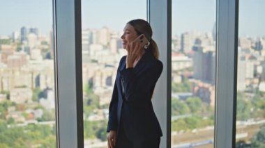 Modern cam ofiste cep telefonuyla konuşan genç bir iş kadını. Gülümseyen işçi panoramik pencerede cep telefonuyla konuşuyor. Takım elbise danışmanlığında rahat, mutlu bir finans uzmanı..