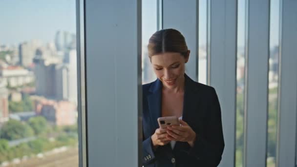 近代的なパノラマオフィスでスマートフォンのタイピングメッセージを読んでいる笑顔の従業員 魅力的なコーポレートマネージャーは 平和的な壊れた携帯電話を楽しんでいます 穏やかな女性ボスは街並みを見ています — ストック動画