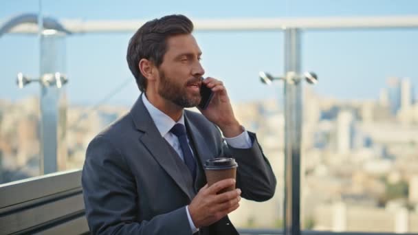 ガラスバルコニーのクローズアップでセルを話すハンサムな労働者 オフィステラスで朝のコーヒーを飲んでいる笑顔の男 成功したビジネスマンは パートナーと良いニュースを共有する携帯電話を話します — ストック動画