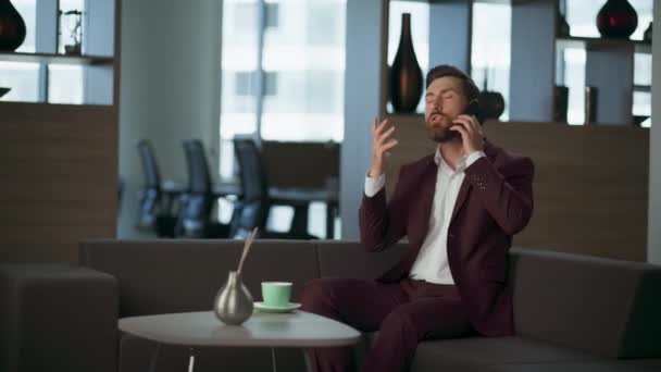 モダンな快適なオフィスのインテリアでスマートフォンを話す怒っているリアクター ストレスのひげの男のプロフェッショナルソファーで呼び出します 大きな窓の近くで計画を議論する集中管理者 コーポレートパーソナルコンセプト — ストック動画