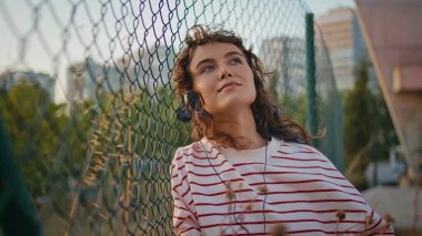 Romantik model, metal tel örgülere yaslanmış müzik dinliyor. Yaz akşamını yakından takip edin. Hayalperest kıvırcık kadın kulaklıklarla güzel melodilerin tadını çıkarıyor. Mutlu genç kız sokakta kulaklık takıyor..