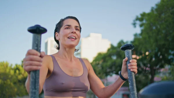 Satisfied lady elliptical machine workout at summer park closeup. Tired sportswoman ending exercises going away from frame. Smiling woman enjoying sport routine loosing weight at open air urban gym