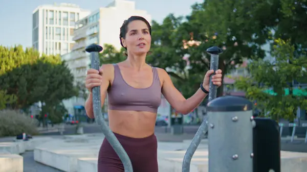 Fitness woman doing elliptical machine exercise at summer park closeup. Satisfied sportswoman ending workout checking results on smartwatch. Smiling lady enjoying sport routine going away from frame
