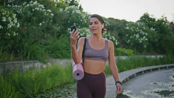 Smiling sportswoman video calling mobile phone strolling green park alley closeup. Calm fit woman walking river embankment talking smartphone videochat on go. Happy fitness lady carrying fit mat