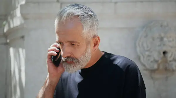 Casual senior talking cell on street closeup. Stylish mature man answering call on travel weekend. Serious grey hair adult discuss business holding phone. Good looking older person with nose piercing.
