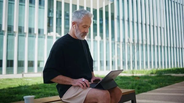 Older manager working remotely in city park. Focused senior typing computer surfing internet in morning. Stylish grey hair entrepreneur using laptop outdoors. Casual bearded man checking data report.
