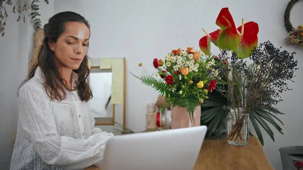 Thoughtful businesswoman type computer laptop in flower store. Focused woman entrepreneur make notes to do list using digital device in floristic shop. Serious girl sit desk in boutique. Work concept.