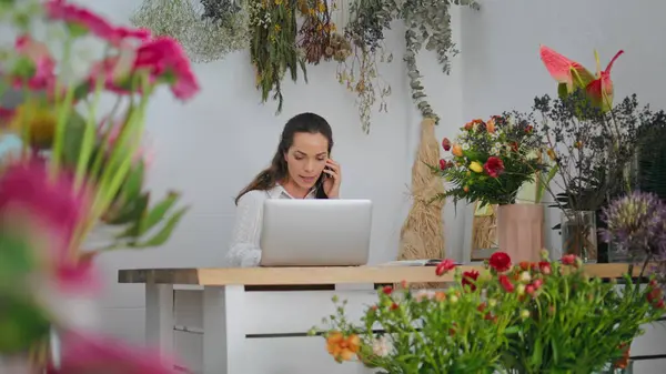 Business florist talk smartphone online in flower shop. Attractive woman work laptop device in plant store workplace. Busy girl type computer in floral boutique. Online communication modern concept.