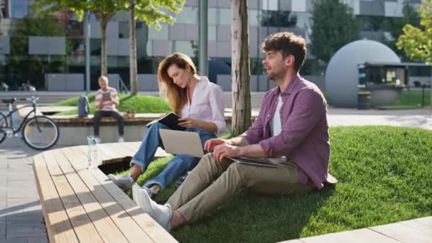 自由撰稿人在城市公园完成工作 微笑的同事在阳光灿烂的街道上遥谈 两个正在学习记笔记的成人学生 松懈的男人合上笔记本电脑聊天迷人的女孩 — 图库视频影像