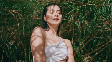 Kaygısız model güneş ışınları içindeki kamera yeşillik portresi. Güneş ışığında poz veren mutlu kız bambu doğasına yakın çekim. Güzel stressiz kadın yaz tatilinde dinleniyor. Haftasonu konsepti 