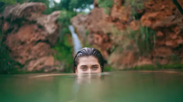 Woman peeping from water surface at wilderness closeup. Wet hair brunette swimming in tropical lagoon with volcanic cliffs background. Relaxed model looking camera enjoying fjord in jungles alone