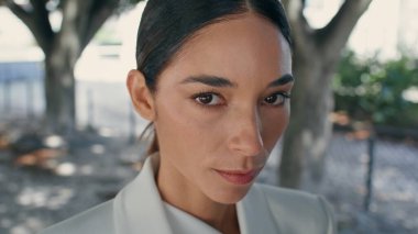 Modayı takip eden beyaz takım elbiseli şık bir iş kadını portresi. Ağaçların arasında poz veren kendine güvenen çekici bir kadın. Yakın plan. Şehir parkında duran göz kamaştırıcı kız. Kadın güzellik konsepti