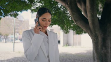 Kadın işyeri sahibi, park ağacının altında dikilirken arıyor. Ciddi, göz kamaştırıcı bir iş kadını dışarıda telefonla konuşuyor. Kendine güveni tam esmer bir kız sokakta akıllı telefondan konuşuyor..