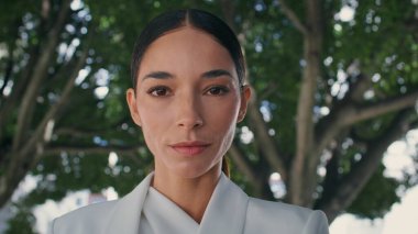 Yeşil ağaçların önünde kameraya bakan lüks bir kadın var. Yaz parkında zarif saç stiliyle poz veren çekici bir iş kadını portresi. Şık iş kadını gülümsüyor..