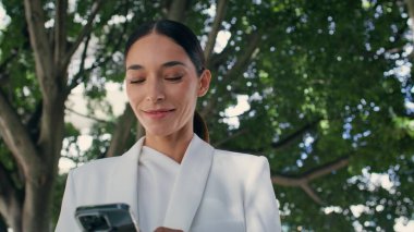 Şehir parkında yürüyen akıllı telefon görünümlü gülümseyen kadın. Çekici başarılı iş kadını dışarıda telefonda iyi haberler okuyor. Güzel esmer kız internette cep telefonuyla sohbet ediyor..