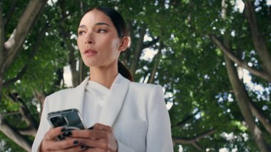 Akıllı telefon görünümlü güzel bir bayan tek başına şehir bahçesinde yürüyor. Modern açık havada telefonla iş yazışmaları yürüten başarılı bir kadın. Kendine güvenen iş kadını telefonu kaydırıyor.