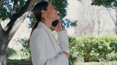 Gülümseyen iş kadını iş yerinde telefonla konuşuyor. Güzel bir parkta yürüyor. Yan görüş çekici, mutlu, esmer, akıllı telefondan konuşan. Göz kamaştırıcı iş kadını telefon konuşmasının keyfini çıkar.