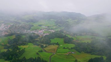 Bitki örtüsü dağlarını kaplayan alçak bulutlar drone görüntüsü. Yeşil yamaçlardaki sisli uzak kır evleri yavaş çekimde. Yağmurlu havalarda nefes kesen yeni tarlalar. Doğa konsepti