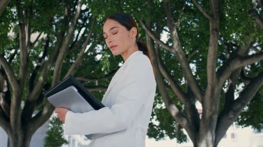 Başarılı bir iş kadını yeşil ağacın önünde kameraya özgüvenle bakıyor. Güzel bir parkta dizüstü bilgisayarla poz veren çekici bir bayan patron. Şık kız gülümsüyor. Kendine güveniyor..