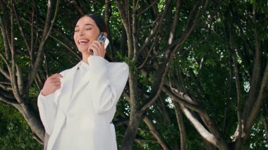 Geniş yeşil ağacın altında telefonla konuşan mutlu iş kadını. Şehir parkında gülen başarılı, zarif bir bayan arkadaşlarını arıyor. Güzel genç bayan telefonda müjdelerle seviniyor..