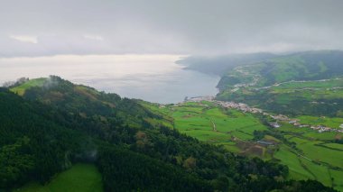 Hava yeşili tepeler bulutlu bir yaz günü. Drone manzaralı dağlık yamaçlar, gök gürültüsü bulutlarının altındaki yemyeşil yeşillikleri kaplıyor. Canlı çimen arazisindeki kırsal köy. Resimli deniz manzarası.