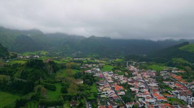 Köy binalarının havadan manzarası. Geniş yeşil vadideki panoramik manzaralı küçük Avrupa evleri. Kırsal yamaçlarda sisli sis yavaş çekimde esiyor. Yağmurlu hava doğası 