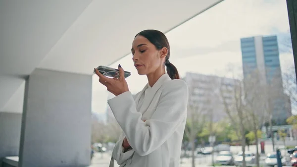 Businesswoman using voice recognition virtual assistant at phone standing at modern building close up. Confident serious woman recording audio message on telephone. Rich lady speaking in loudspeaker.