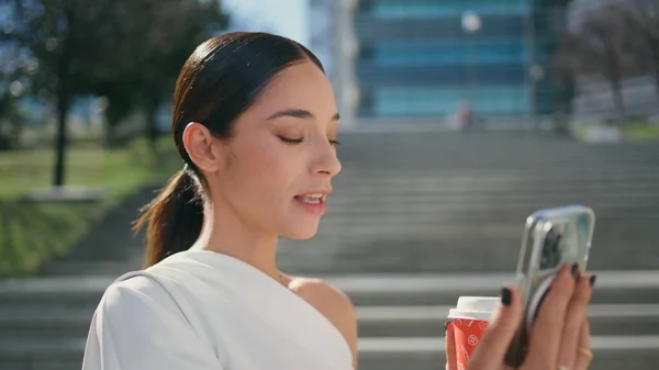 Joyful woman video calling at smartphone standing sunny city staircase close up. Happy smiling girl drinking coffee looking telephone web camera. Cheerful brunette recording life video blog outdoors.