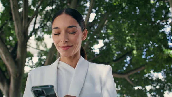 Şehir parkında yürüyen akıllı telefon görünümlü gülümseyen kadın. Çekici başarılı iş kadını dışarıda telefonda iyi haberler okuyor. Güzel esmer kız internette cep telefonuyla sohbet ediyor..