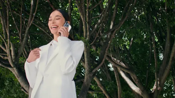 Geniş yeşil ağacın altında telefonla konuşan mutlu iş kadını. Şehir parkında gülen başarılı, zarif bir bayan arkadaşlarını arıyor. Güzel genç bayan telefonda müjdelerle seviniyor..