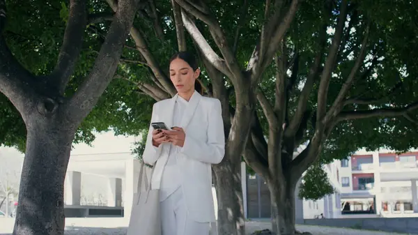 Businesswoman dial phone number walking under beautiful green trees. Elegant chic business lady in white suit calling telephone outdoors. Serious gorgeous woman using smartphone for work communication