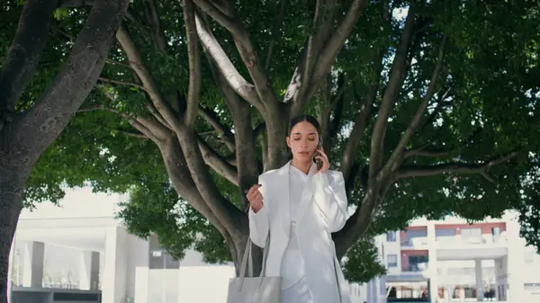 Zengin bir kadın zarif beyaz takım elbisesiyle akıllı telefon park yolunu arıyor. Başarılı iş kadını yeşil ağaçların altında telefonla konuşuyor. Gülümseyen lüks kız telefonda konuşuyor..