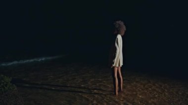 Kız silueti gece sahilinde deniz suyuna basıyor. Zayıf, yalnız bir kadın okyanus kıyısında yürüyordu. Beyaz ceketle koyu arka planda rüya görüyordu. Tanımlanamayan Afro-Amerikalı ışıkta sığ dalgalar yaratıyor.. 