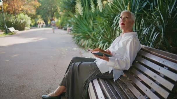 シティパークで本を読んでいる上級女性 朝のベンチに座っている緑の庭を賞賛する美しい灰色の髪の女性 文学を屋外で休むメイクを施した平和なスタイリッシュな年金 — ストック動画