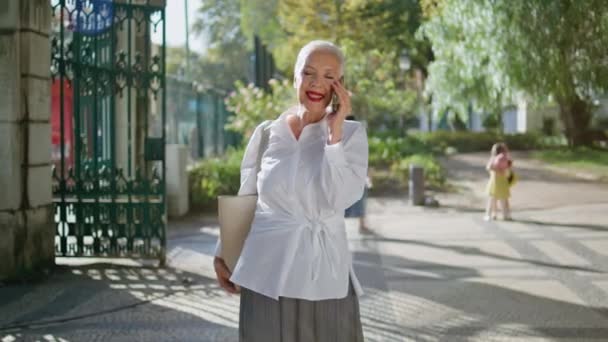 シティガーデンで携帯電話を話すハッピー年金者 笑顔のシニアは 日当たりの良い通りの通話会話を楽しんでいます エレガントな灰色の髪のビジネスマンは ニュースを共有する友人 退職生活スタイルコンセプト — ストック動画