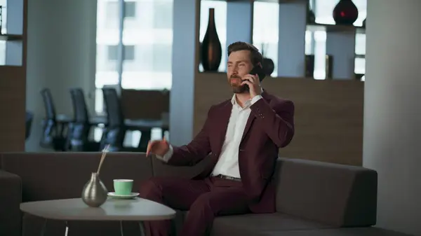 Joyful coach talking cellphone in office interior. Successful man manager laughing at sofa modern hall. Cheerful businessman working project mobile phone in open space. Financial discussion concept.